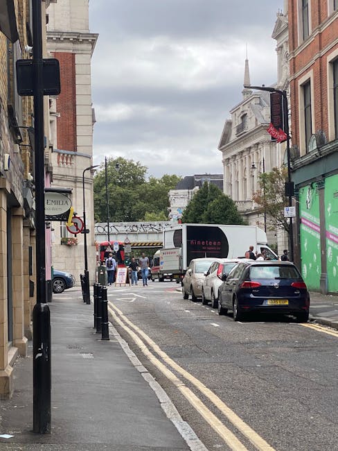 A narrow urban street in Putney during daytime, showing parked cars along the curb, a white moving truck with a closed cargo area positioned at the street corner, and a few people visible near the tru
