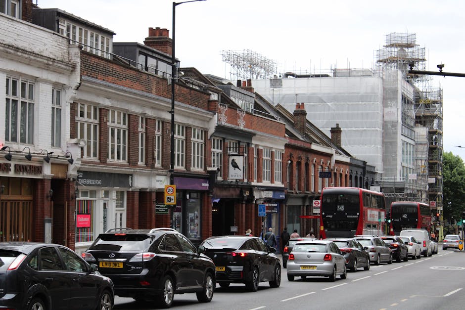 A narrow urban street in Putney during daytime, showing parked cars along the curb, a white moving truck with a closed cargo area positioned at the street corner, and a few people visible near the tru