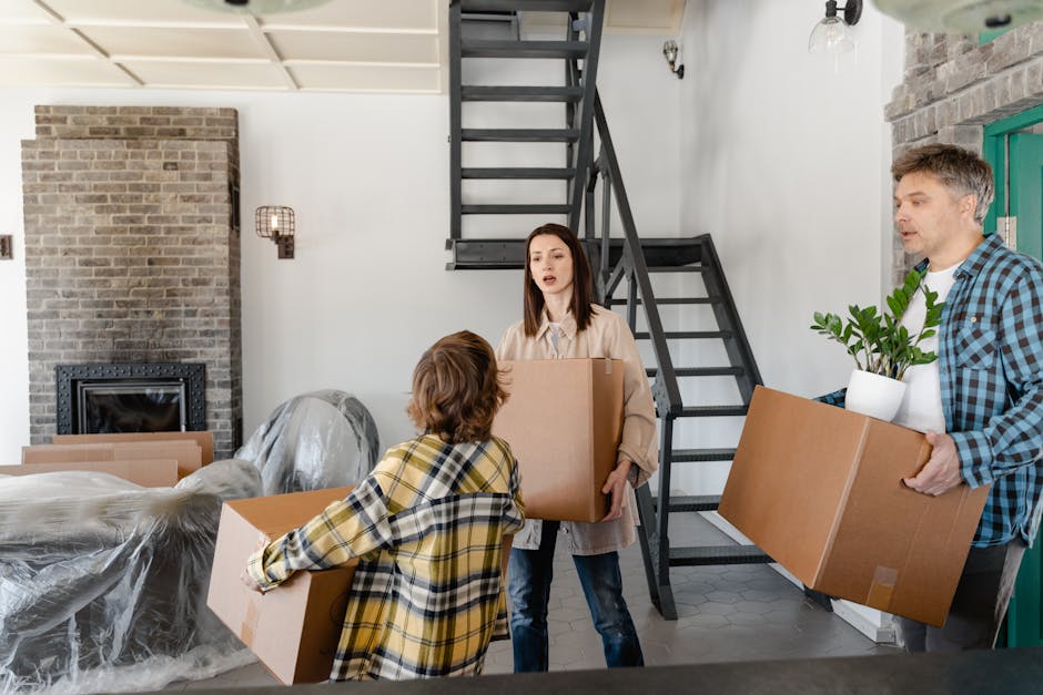 A man and a woman lie on the floor inside a residential property surrounded by packed cardboard boxes, some sealed with packing tape and others open, revealing household items. The woman has long brow