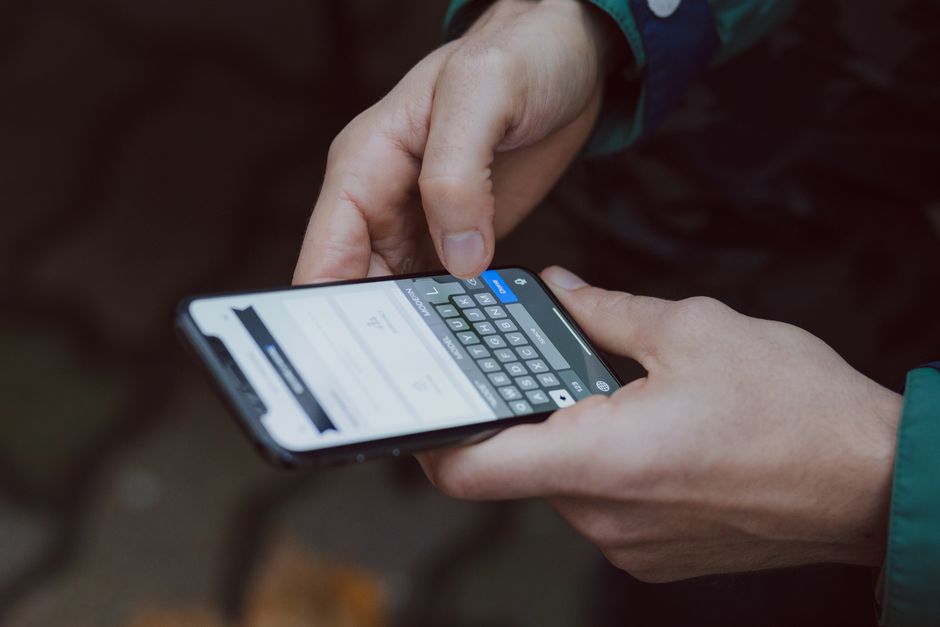 Close-up of a person's hands holding a smartphone, typing on the device's virtual keyboard with the screen displaying a form or message. The individual is wearing a dark jacket or coat, and the backgr