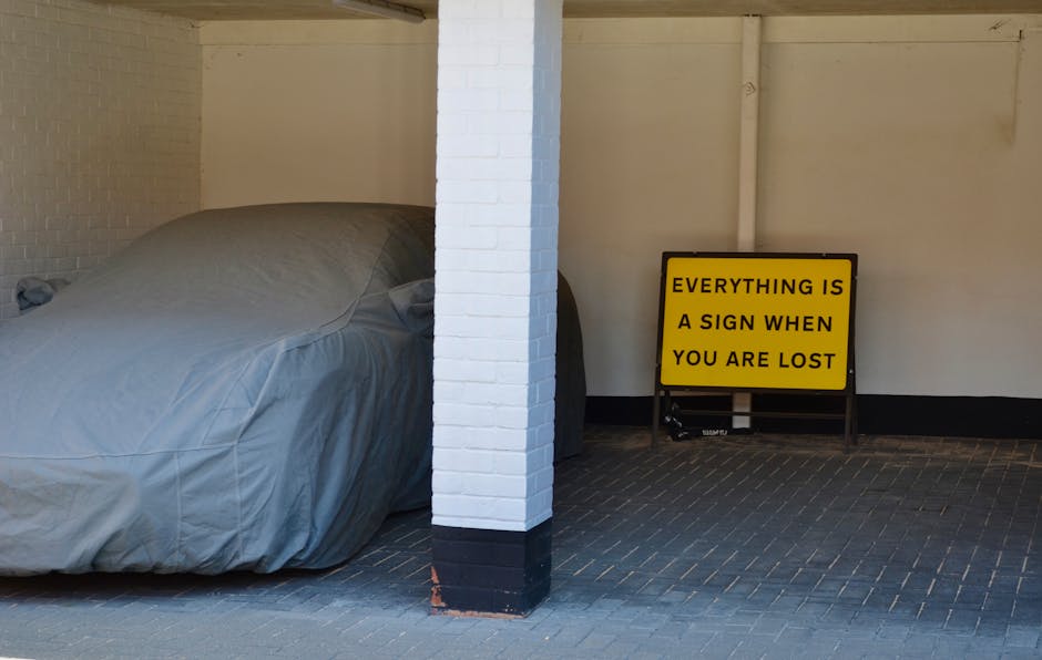 A covered vehicle, possibly a car or small van, parked indoors next to a white-painted brick pillar, with a yellow sign on a stand nearby that reads 'EVERYTHING IS A SIGN WHEN YOU ARE LOST.' The vehic