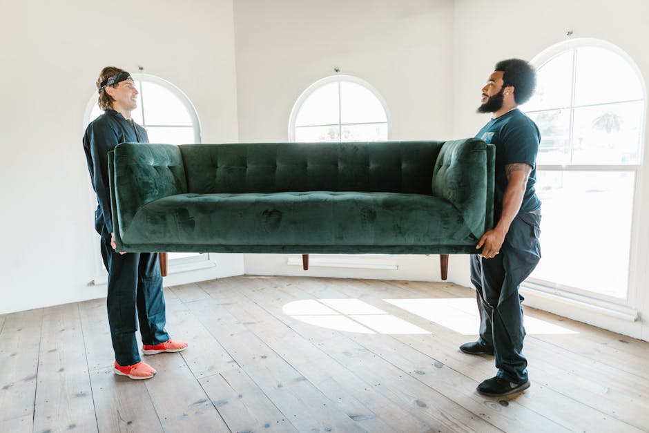 Two movers from Putney Removals are carefully carrying a large, dark green velvet sofa through a spacious, well-lit room with light wooden flooring. The sofa appears to be a three-seater with a tufted
