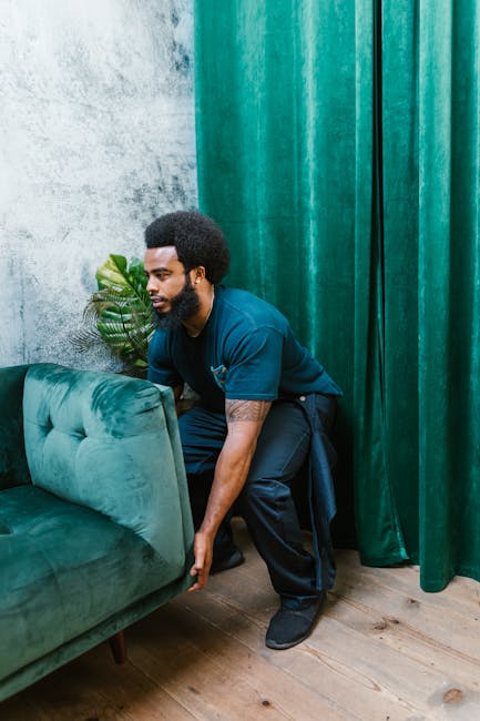 Two movers from Putney Removals are carefully carrying a large, dark green velvet sofa through a spacious, well-lit room with light wooden flooring. The sofa appears to be a three-seater with a tufted