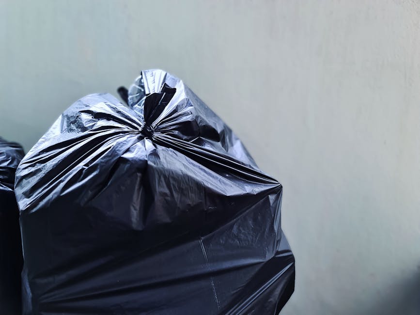A person wearing a red shirt and beige pants is seen during a home relocation process, placing a clear plastic container with a lid next to a white plastic recycling bin labeled 'PLASTIC,' inside a ro