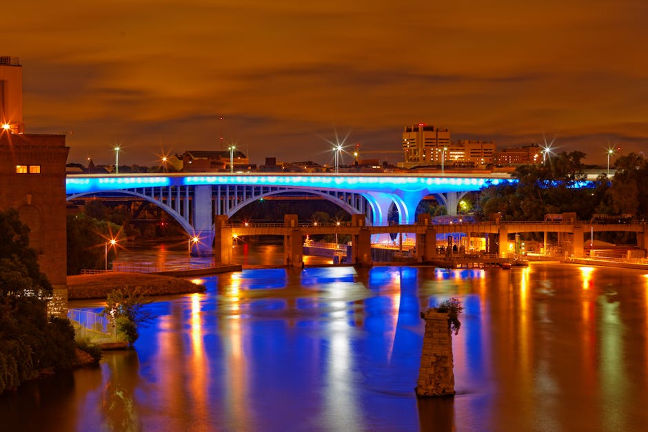 A nighttime view of a city bridge illuminated with blue and white lights, spanning over a calm river that reflects the colorful lighting. The bridge features arched supports and is lined with streetli