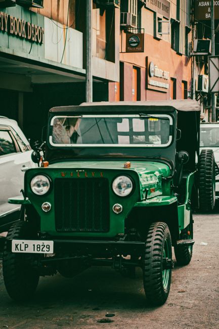 A vintage green utility vehicle with a boxy front and round headlights is parked on a street in an urban area. The vehicle features a black grille and windshield, with a metal roof supported by visibl