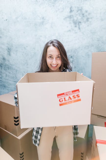 A young woman with long brown hair and a cheerful expression is holding an empty cardboard box labeled 'fragile glass' in a room with a light blue textured wall. She is wearing a casual plaid shirt an