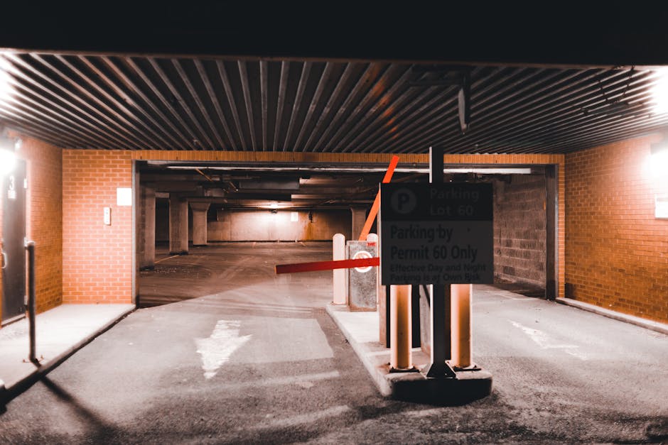 The image shows the entrance to an underground parking garage with a lowered security barrier arm made of orange plastic, positioned across the driveway. In front of the barrier, there is a parking re